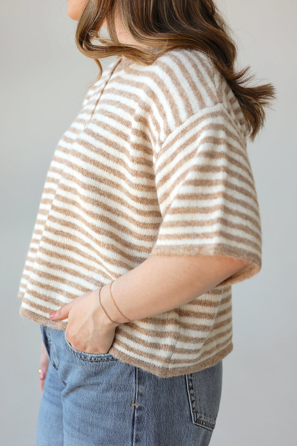 A woman shown from the side wearing a fuzzy short-sleeve knit top with horizontal beige and off-white stripes, paired with blue jeans, against a plain light gray background.