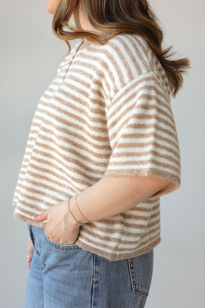 A woman shown from the side wearing a fuzzy short-sleeve knit top with horizontal beige and off-white stripes, paired with blue jeans, against a plain light gray background.