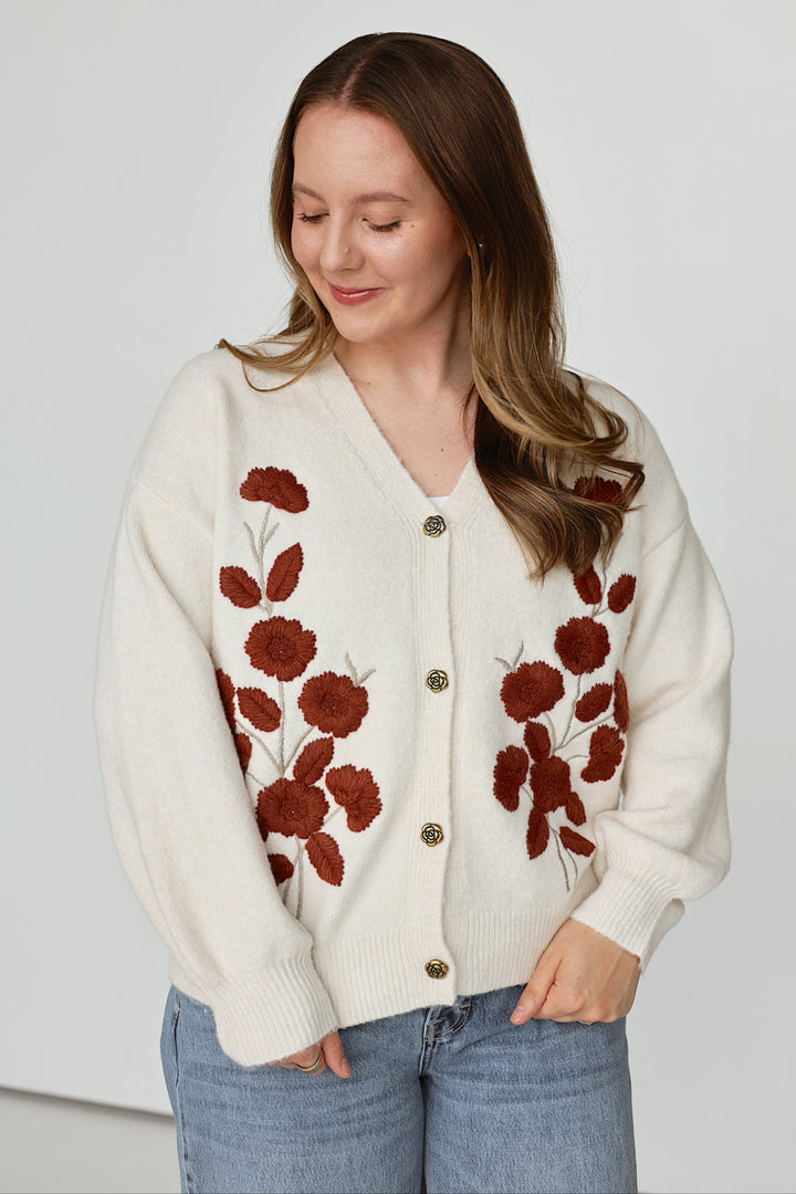 A woman wearing a cream-colored knit cardigan with brown floral embroidery and gold buttons, paired with blue jeans, standing against a plain light gray background.