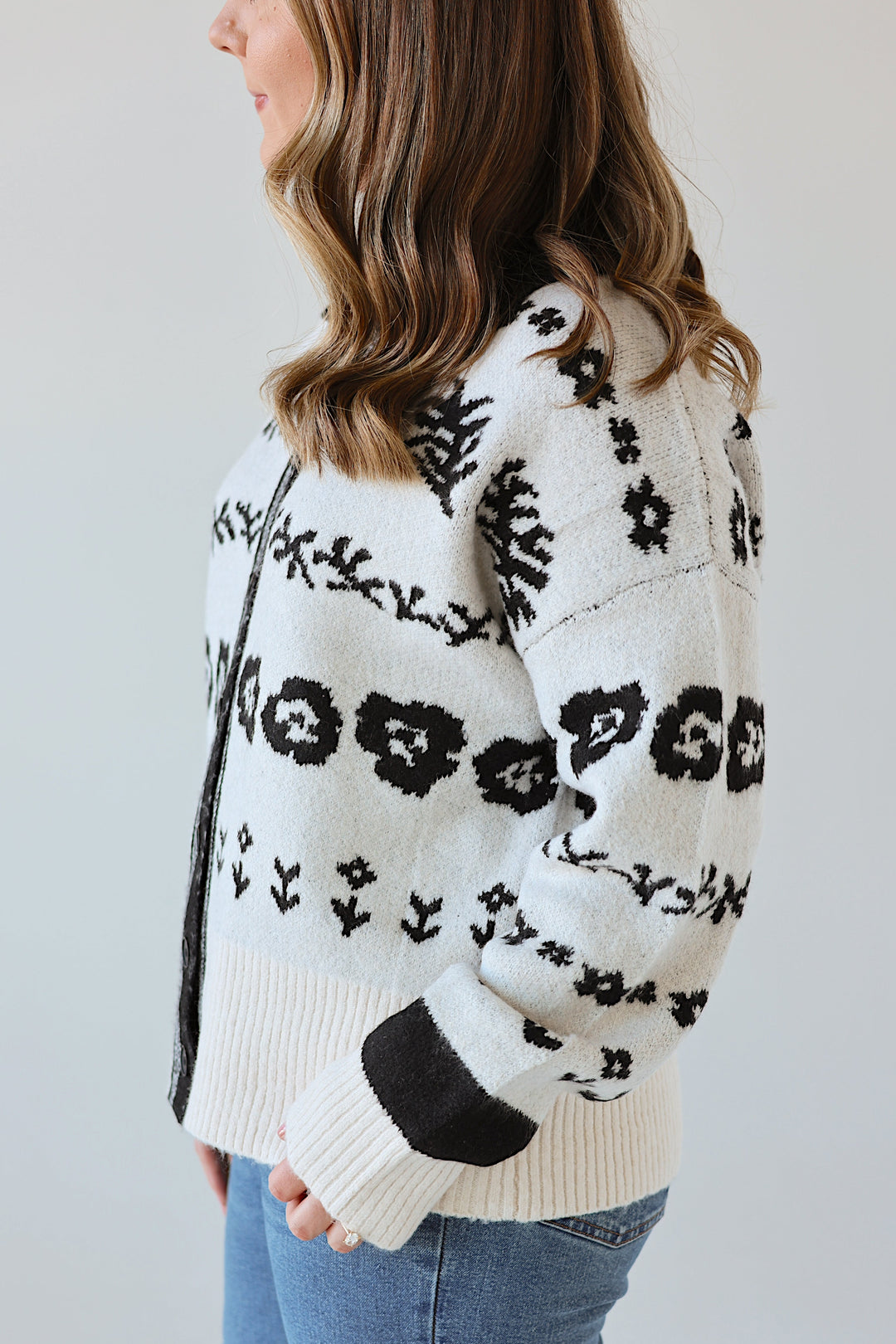 Person wearing a white sweater with black floral patterns and blue jeans on a plain background