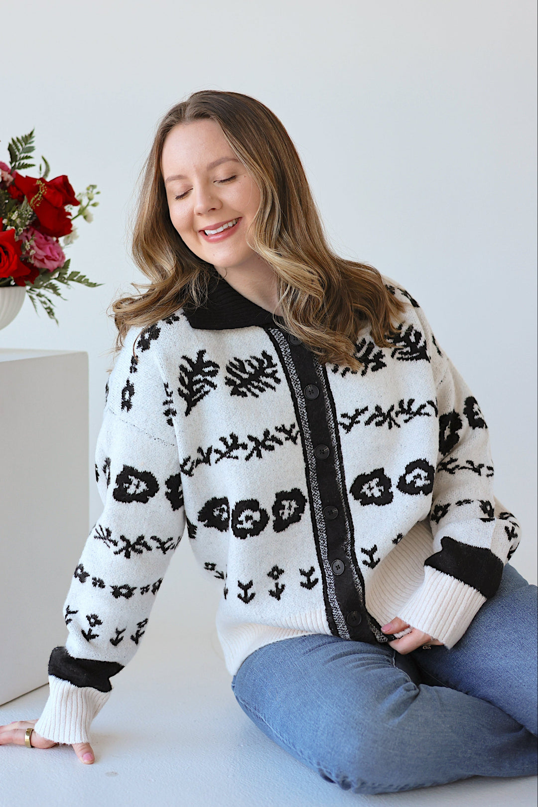 Woman wearing a patterned cardigan sitting next to a vase of flowers on a white background
