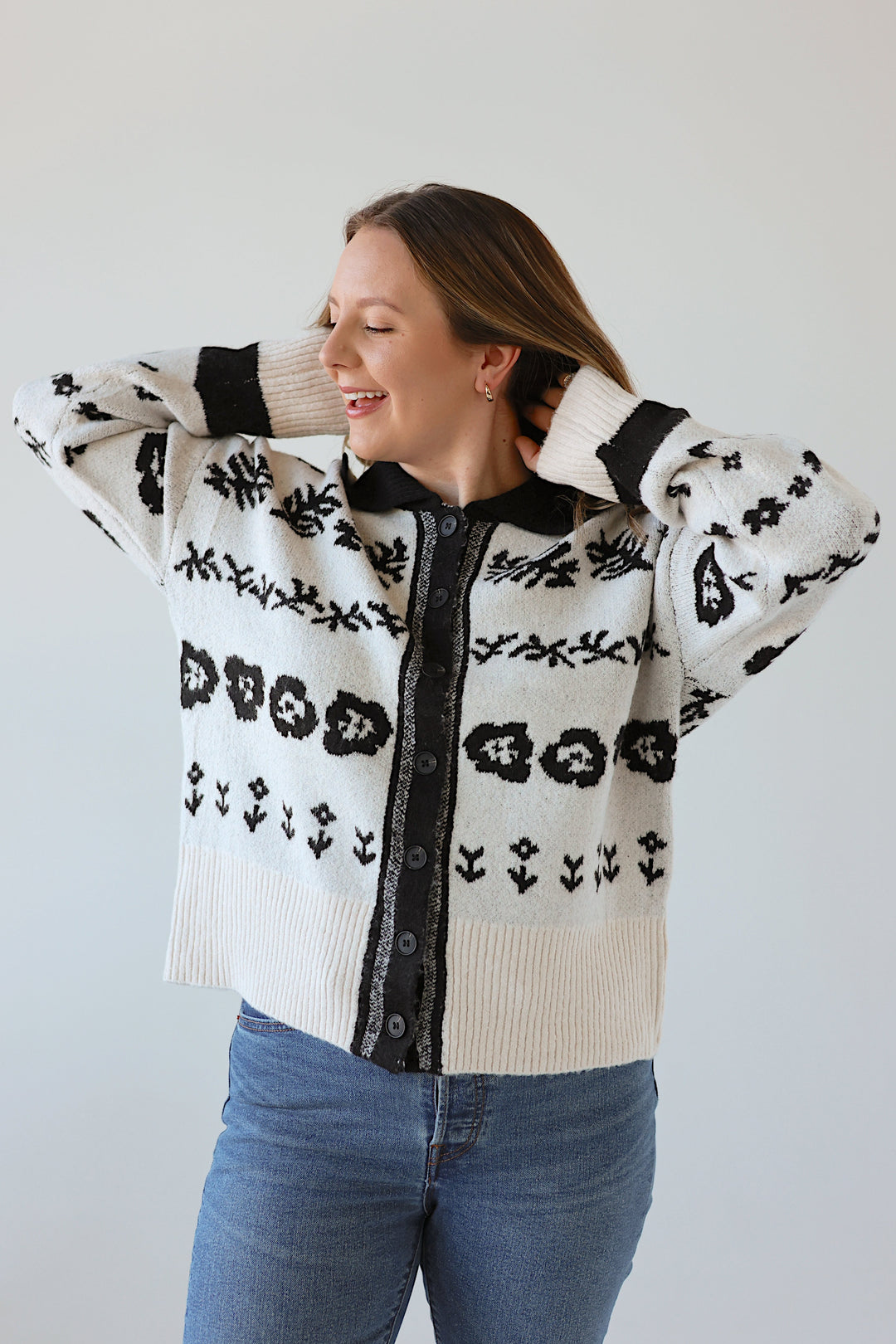 Woman wearing a white sweater with black floral patterns and blue jeans against a plain background