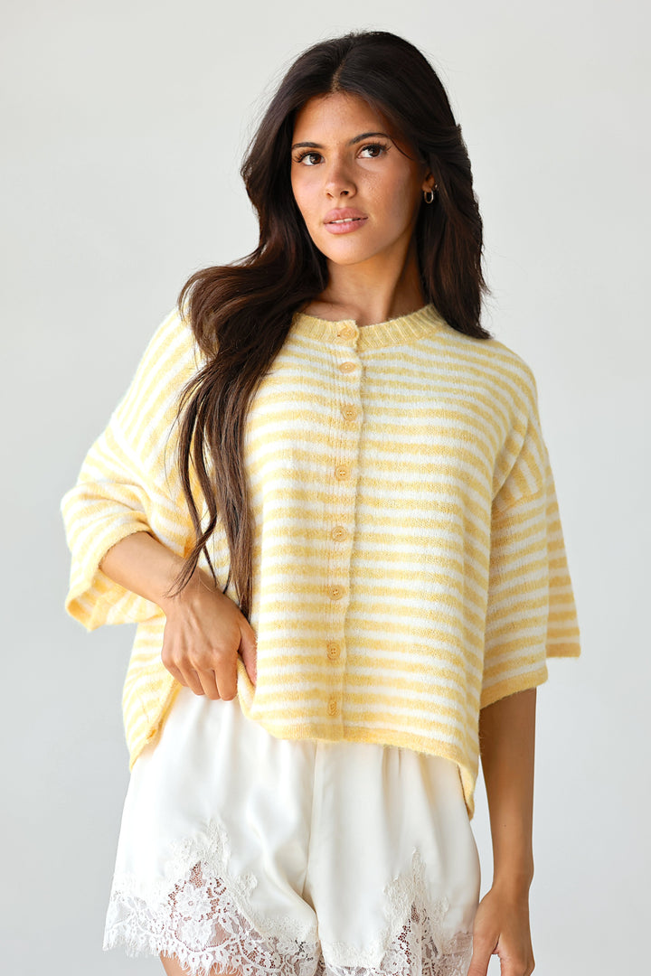 A woman wearing a yellow and white striped short-sleeve top and white shorts with lace trim, photographed against a plain light gray background.