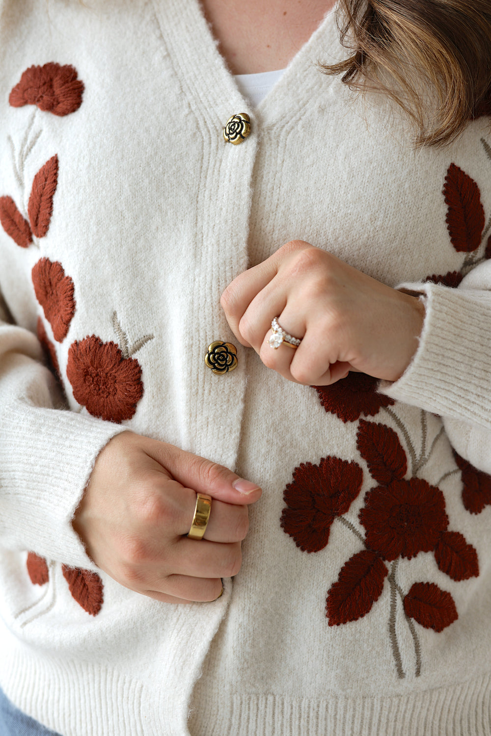 A close-up of a person wearing a cream-colored knit cardigan with dark brown floral embroidery and gold-tone buttons. The person's hands are visible adjusting the front of the cardigan, wearing gold and silver rings.