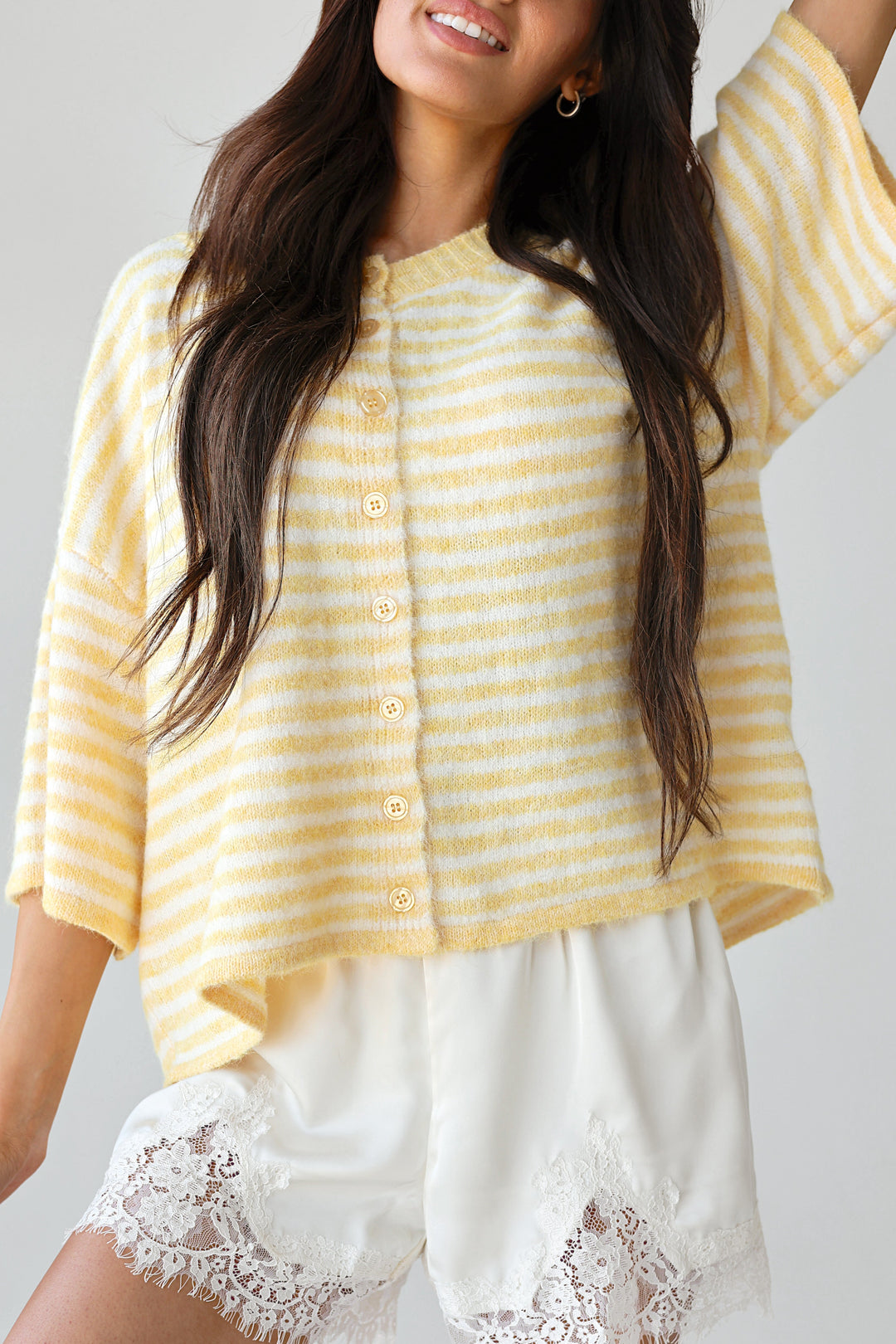 A woman wearing a yellow and white striped short-sleeve button-front top with white shorts featuring lace trim at the hem, posed against a light gray studio background.