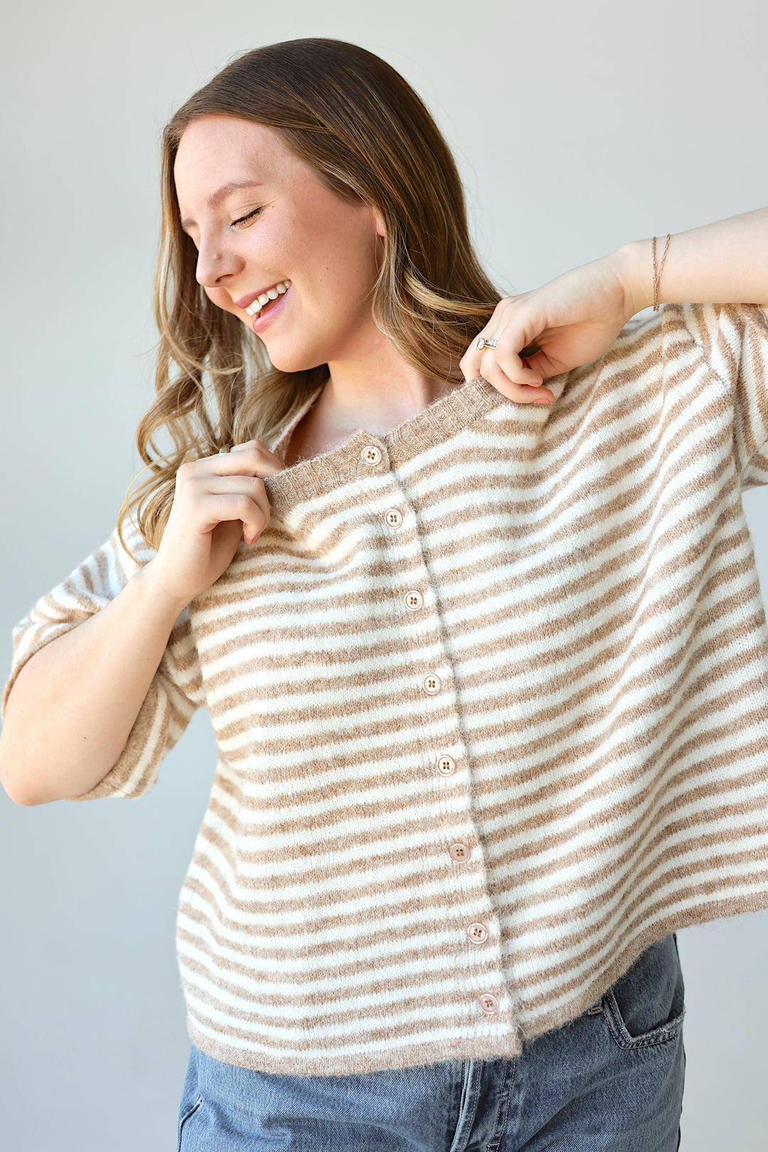 A woman wearing a beige and white striped knit cardigan with a button-up front, posing to show the texture and fit of the garment against a plain light gray background.