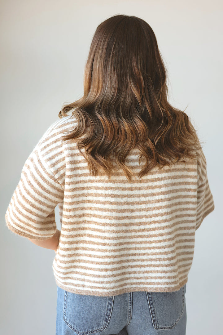 A woman viewed from behind wearing a cream and tan striped short-sleeve knit top and light blue jeans against a plain light gray background.