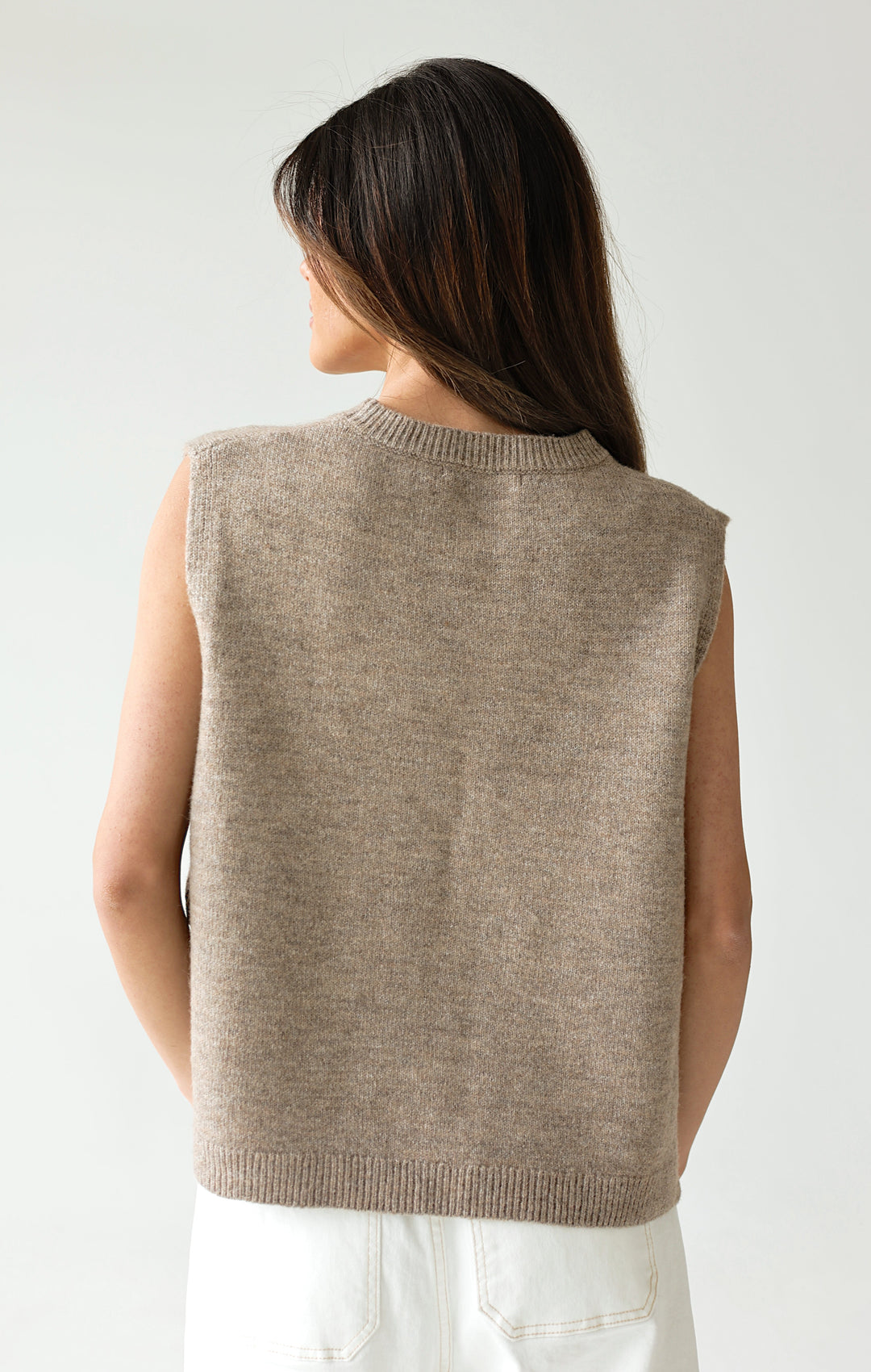 Woman wearing a beige sweater over white pants on a plain background
