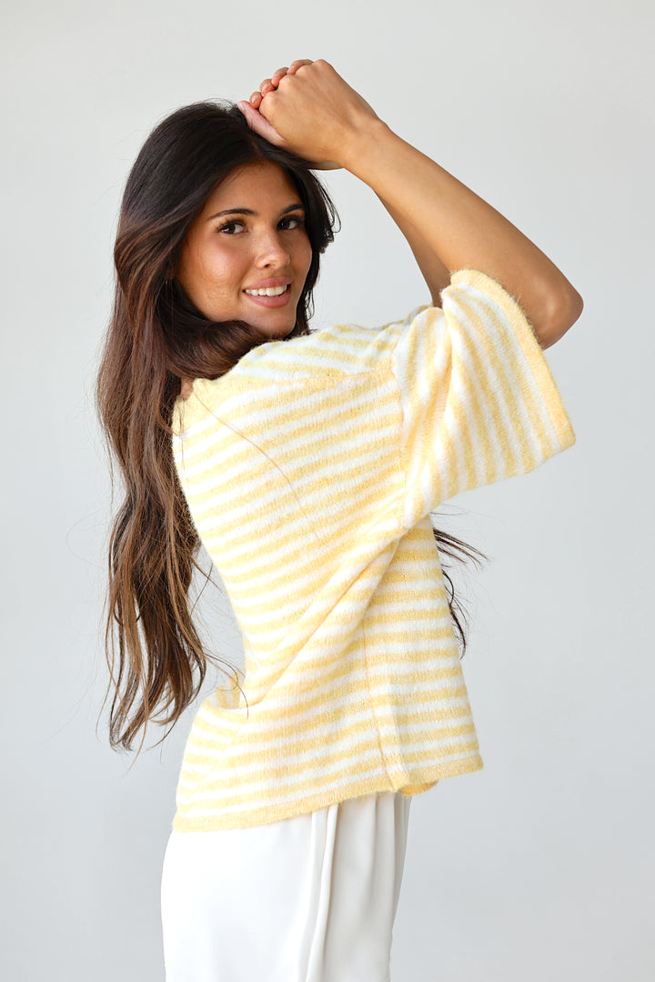 A woman modeling a yellow and white striped short-sleeve button-up top with white lace-trimmed shorts, standing against a light gray studio background.