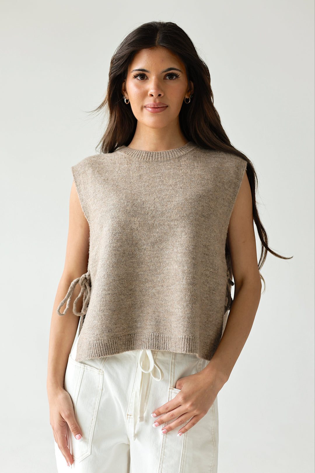 Woman wearing a beige sweater over white pants on a plain background