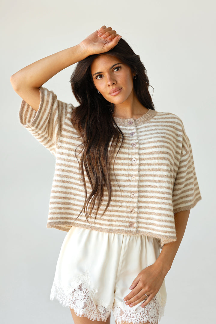 A woman wearing a beige and white striped short-sleeve top and white lace-trimmed shorts, posed against a plain light gray background.