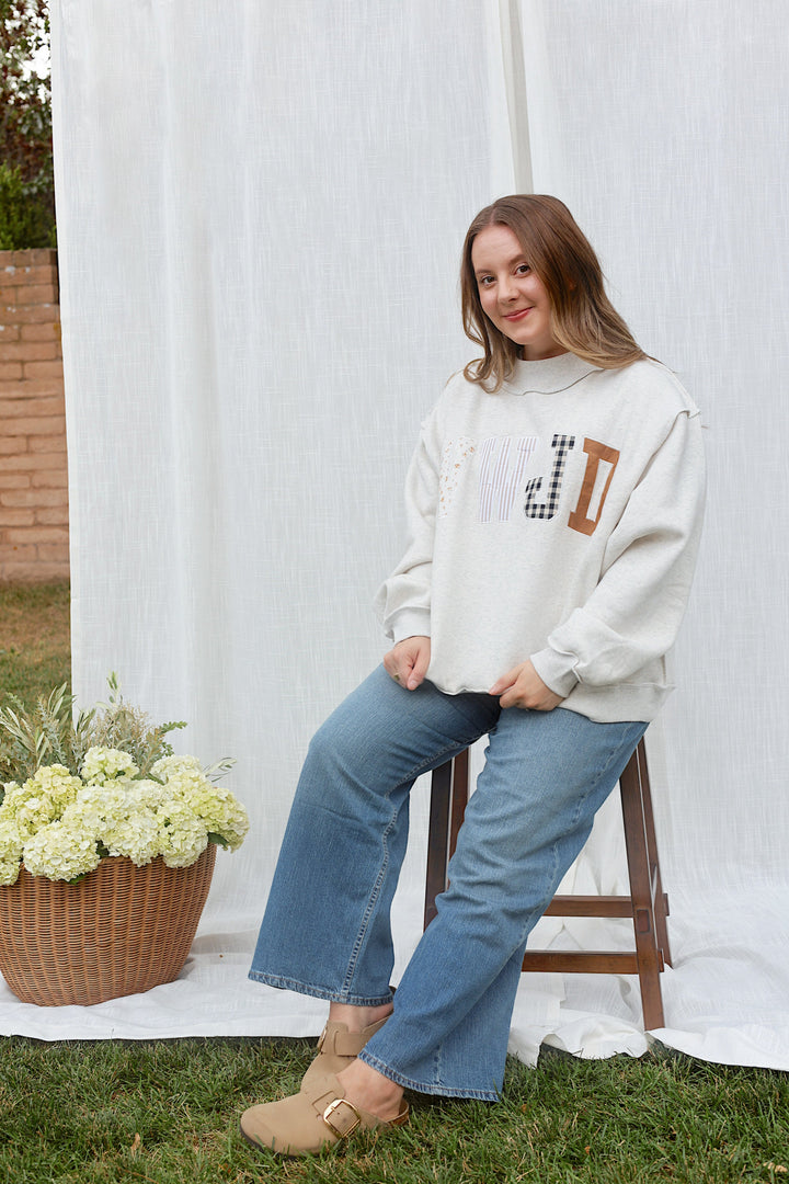 Model wearing the ash grey JESUS embroidered mockneck sweatshirt with jeans, seated full-length view.