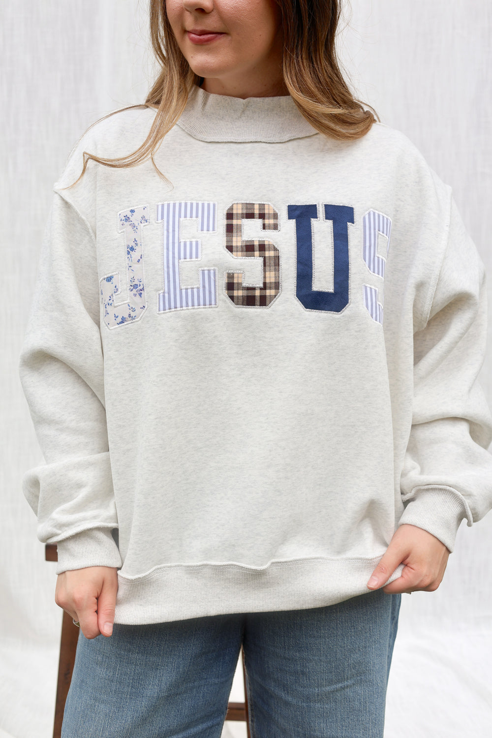 A woman wearing a light heather gray sweatshirt with 'JESUS' printed across the chest in blue, brown, and striped lettering, paired with blue jeans.