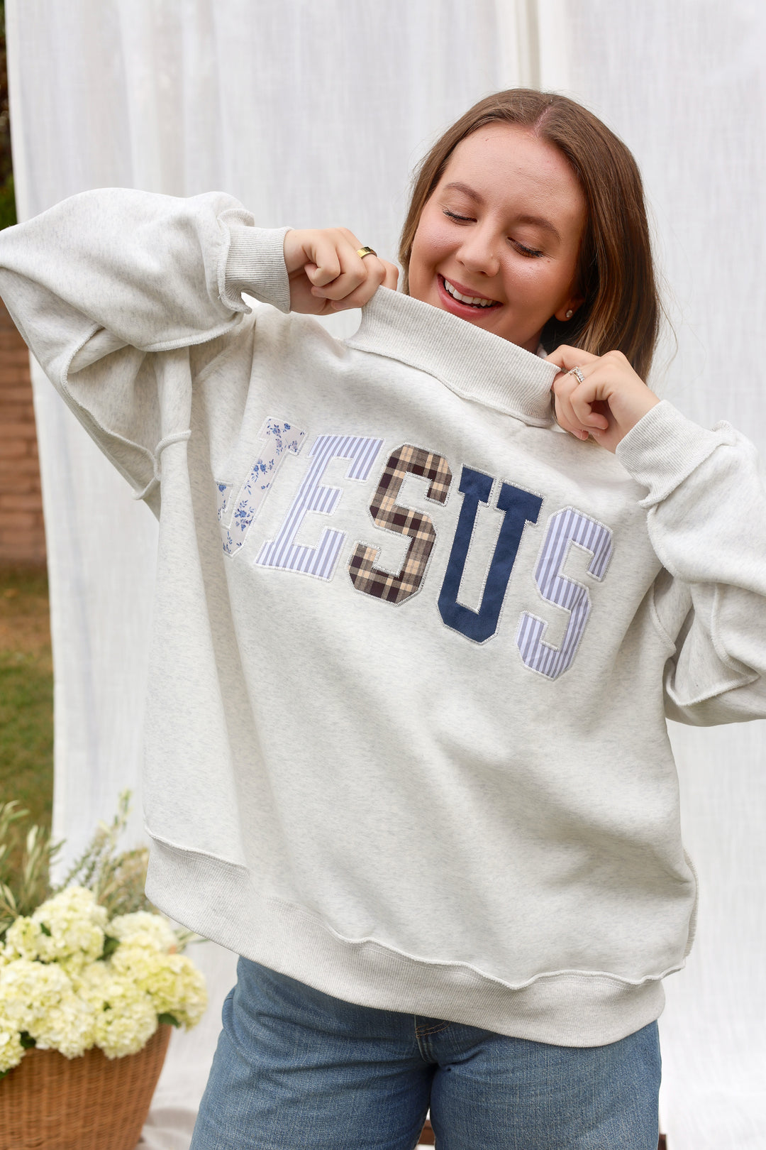 A woman wearing a light beige crewneck sweatshirt with 'JESUS' written across the chest in blue, navy, and plaid lettering, paired with blue jeans.