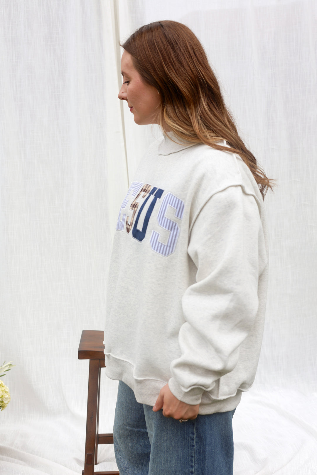 A woman wearing a cream-colored sweatshirt with a blue and white striped graphic on the front, paired with blue jeans, standing in front of a white fabric backdrop.