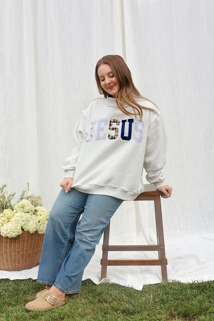 A woman wearing a cream-colored sweatshirt with a graphic patch on the chest, blue jeans, and tan loafers sits on a wooden stool in front of a white fabric backdrop on a grassy lawn.