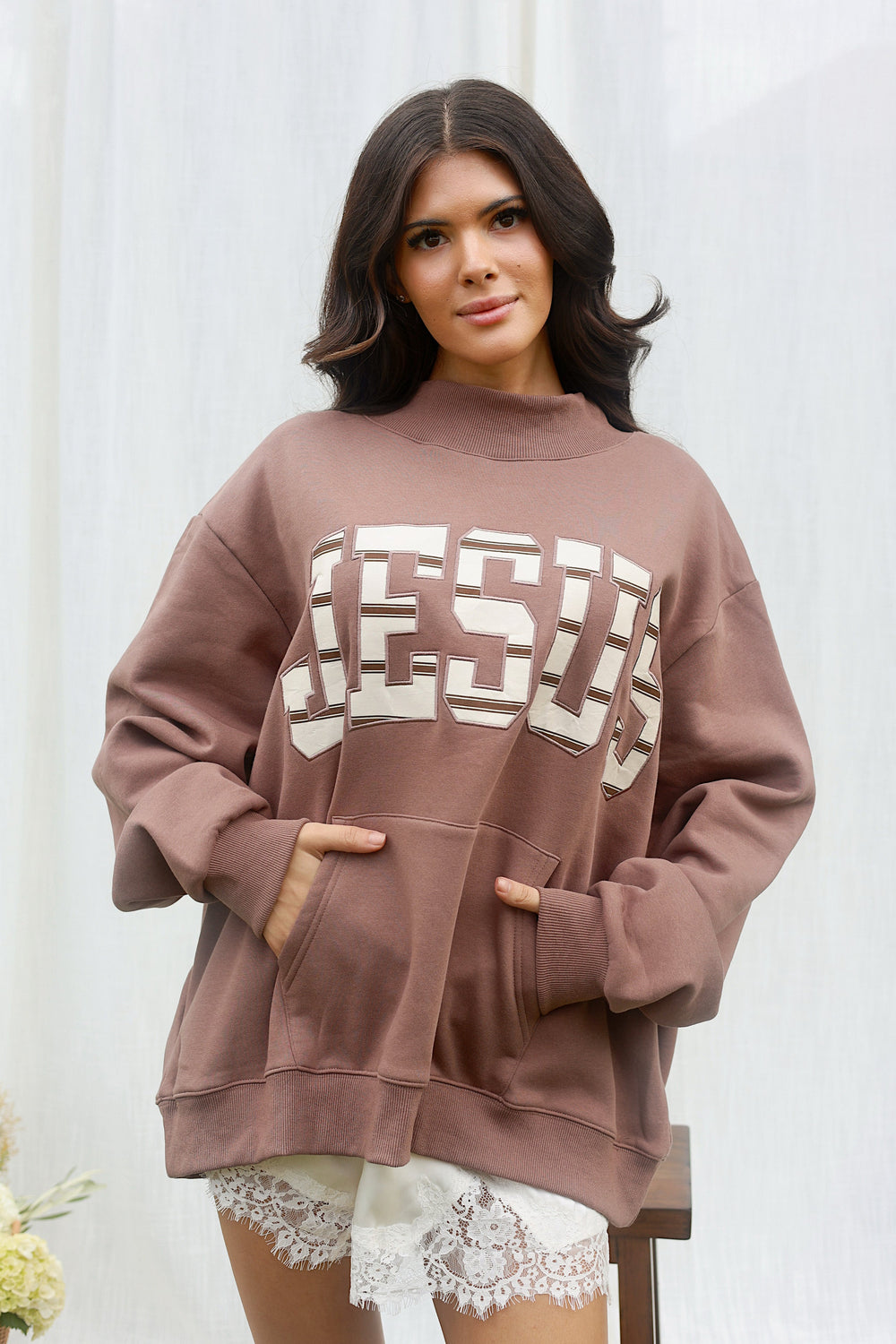 A woman wearing an oversized brown sweatshirt with 'JESUS' printed across the chest in large white block letters, paired with white lace shorts, posed against a white curtain background.