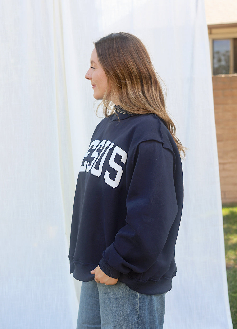Side view of model wearing the oversized navy JESUS embroidered mockneck sweatshirt with jeans.