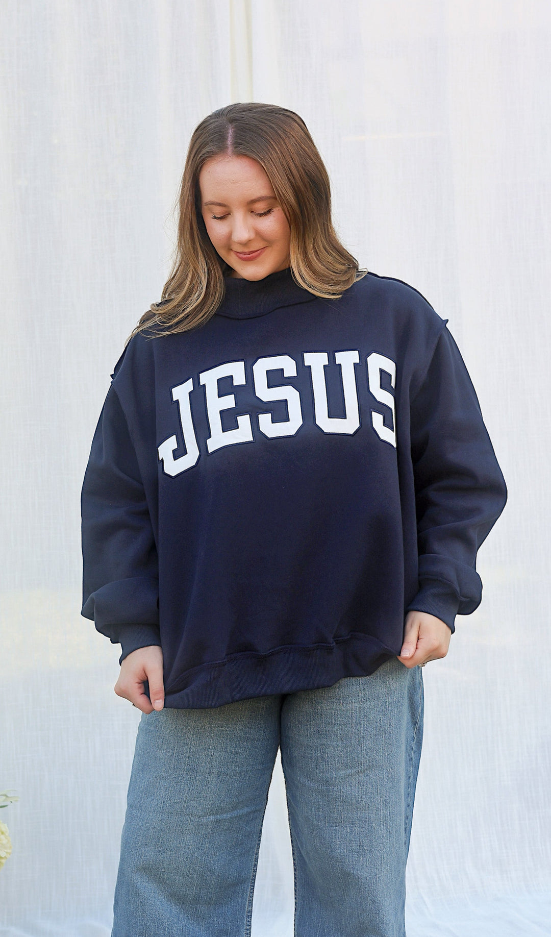 A woman wearing a navy blue sweatshirt with 'JESUS' printed in white varsity letters across the chest, paired with wide-leg blue jeans and tan shoes, standing outdoors in front of a white draped backdrop.